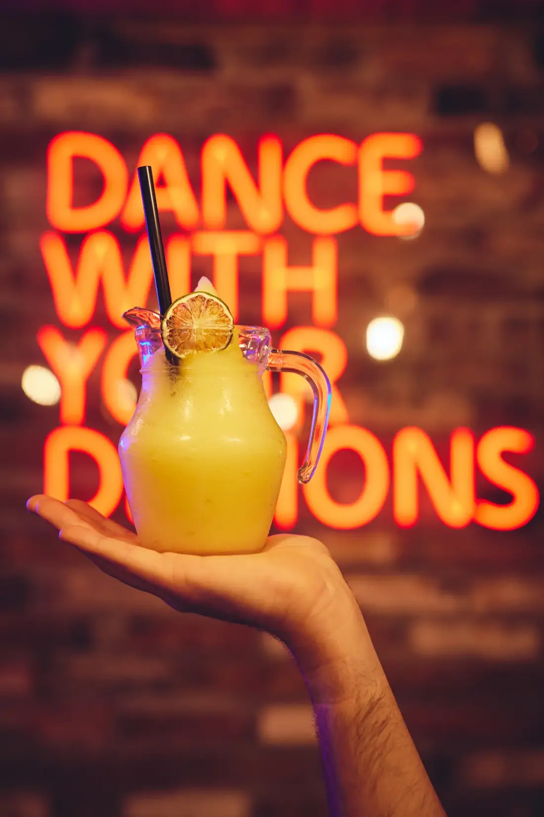 Frozen pitcher cocktail held up in front of the 'Dance with your demons' neon sign